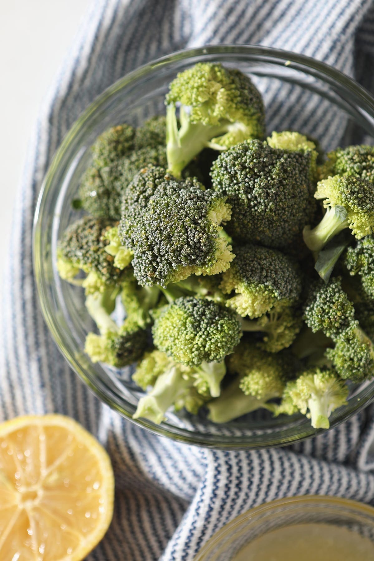 A clear glass bowl holds fresh chopped broccoli on a blue and white striped towel next to a lemon wedge