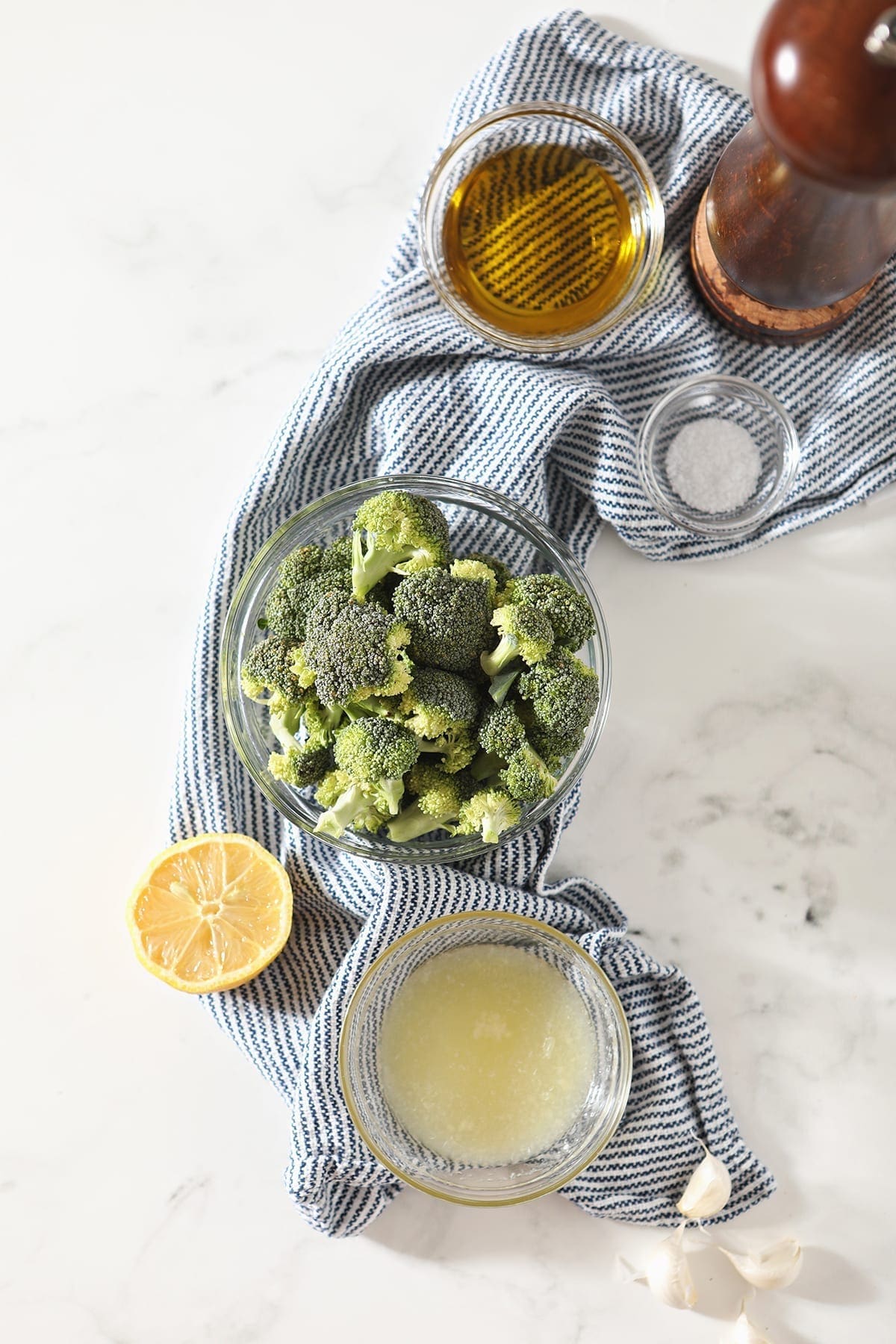 Broccoli, lemon juice, salt, pepper and oil are shown in bowls on a blue and white striped towel on marble