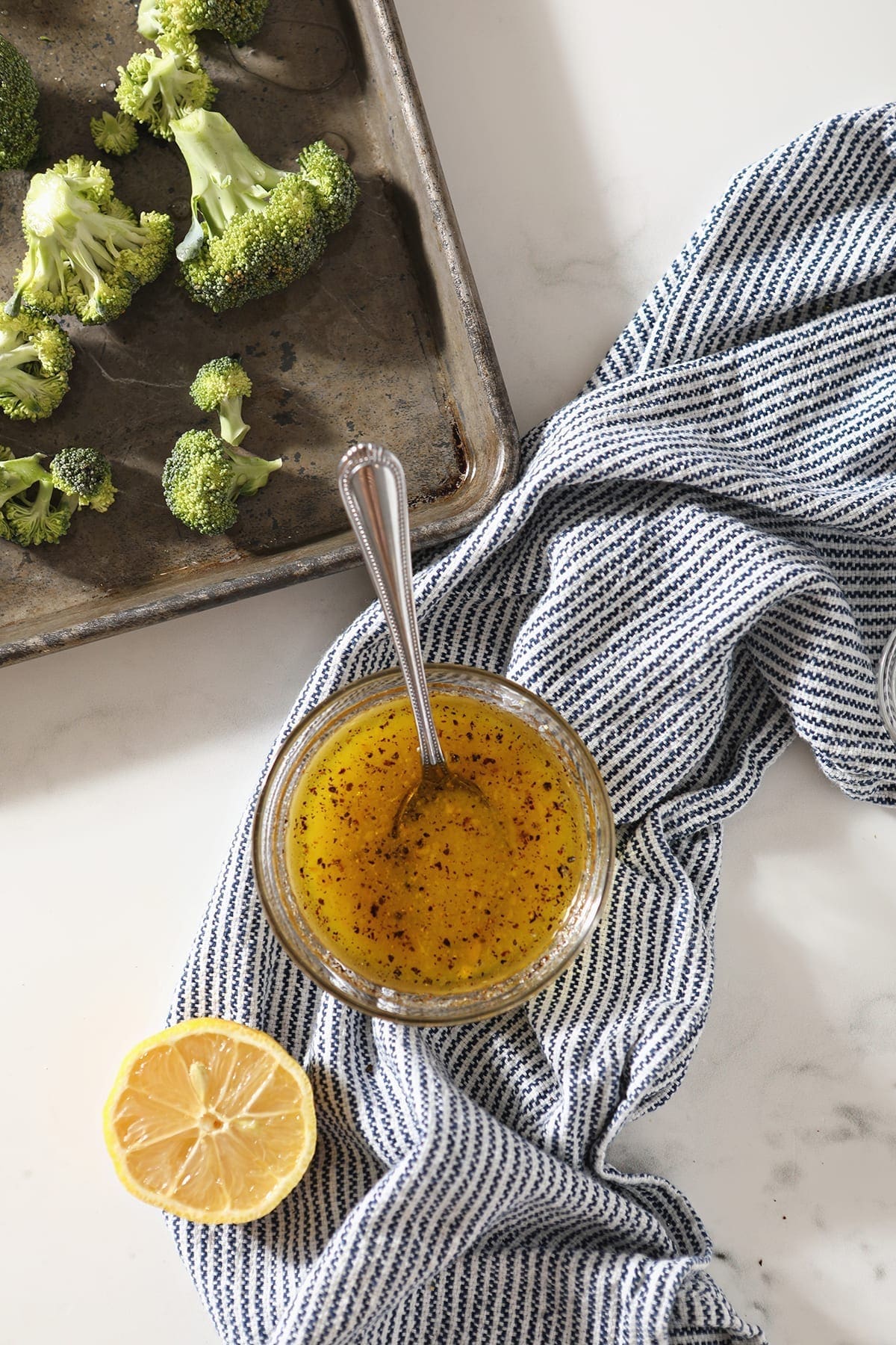 A vinaigrette in a glass bowl sits next to a lemon wedge and broccoli on a sheet pan