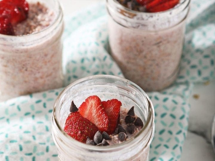 Four containers of strawberry overnight oats sit on a turquoise kitchen towel, garnished with strawberries and other ingredients
