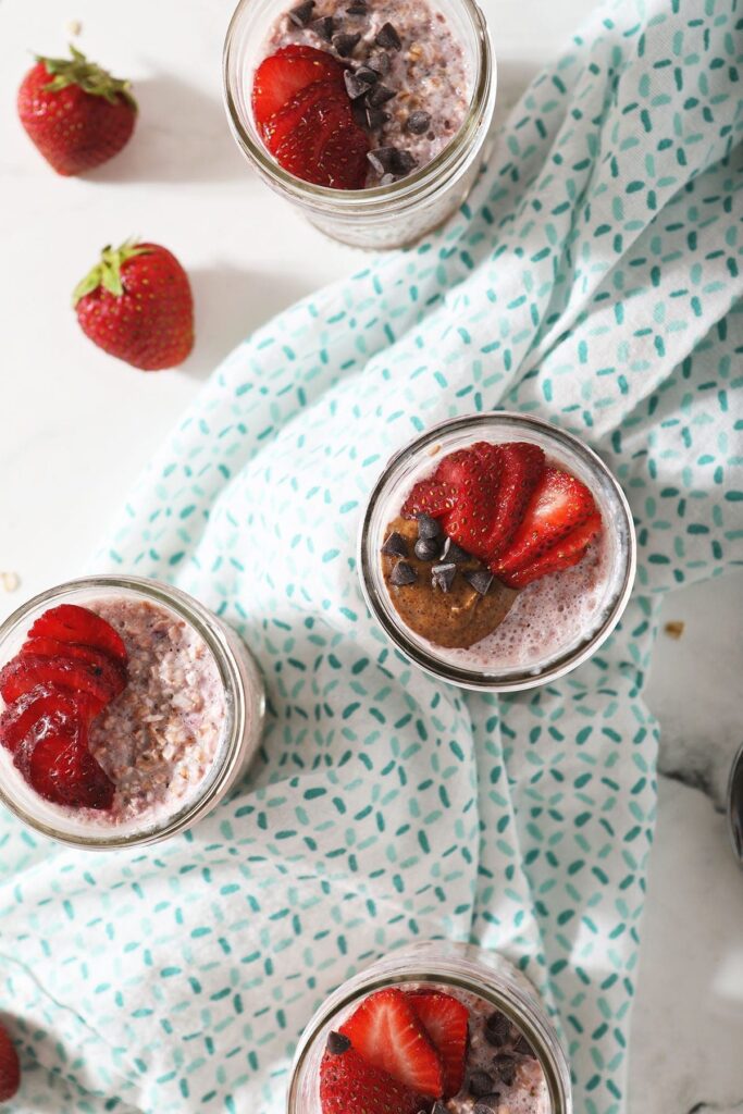 Four servings of strawberry overnight oats in mason jars, garnished with strawberries, almond butter and chocolate chips