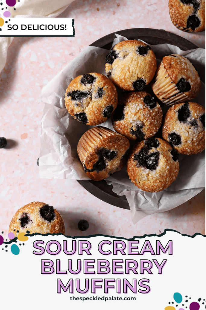 A bowl of Blueberry Muffins in a bowl on a pink surface surrounded by more muffins, orange wheels and blueberries with the text sour cream blueberry muffins