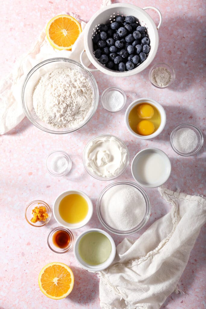 Ingredients to make Sour Cream Blueberry Muffins with Orange in bowls on a pink surface with a beige linen