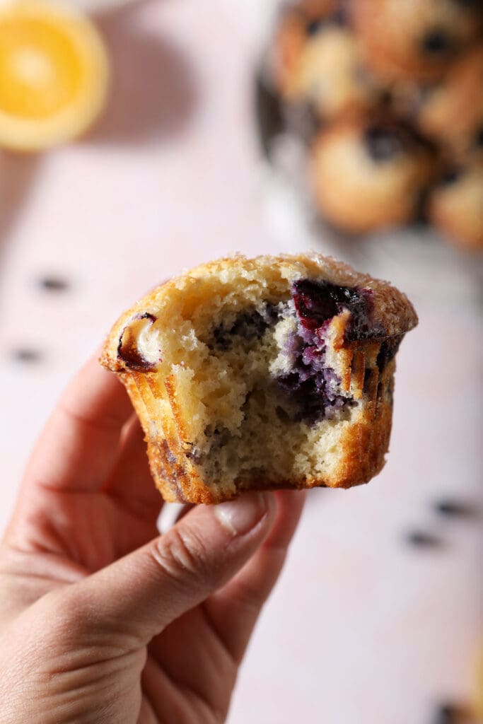 A hand holds a bitten-into Sour Cream Blueberry Muffin above a pink surface with a plate of muffins and an orange