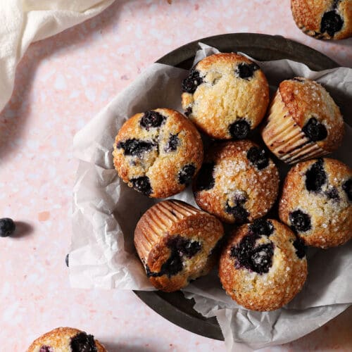 A bowl of Blueberry Sour Cream Muffins in a bowl on a pink surface surrounded by more muffins, orange wheels and blueberries