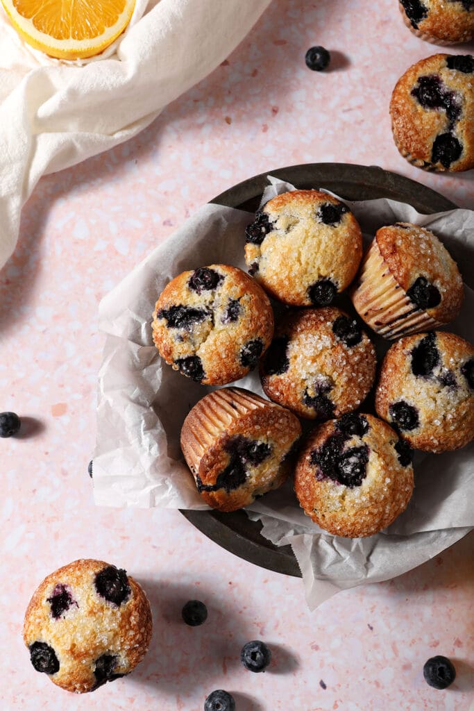 A bowl of Blueberry Sour Cream Muffins in a bowl on a pink surface surrounded by more muffins, orange wheels and blueberries