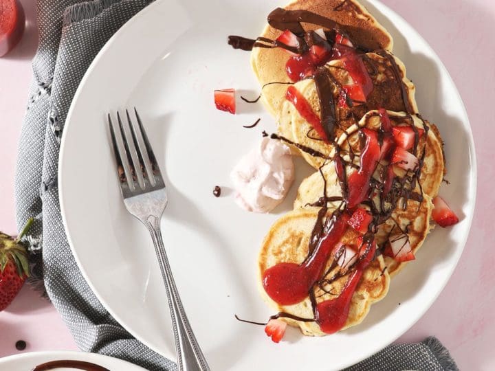 Three white plates holding Strawberry Pancakes with whipped cream, dark chocolate and strawberry sauce