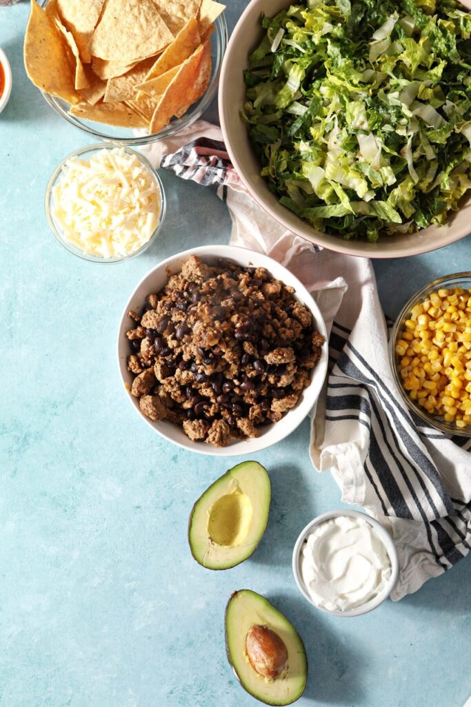 Black beans and ground turkey in a white bowl surrounded by taco salad ingredients in bowls