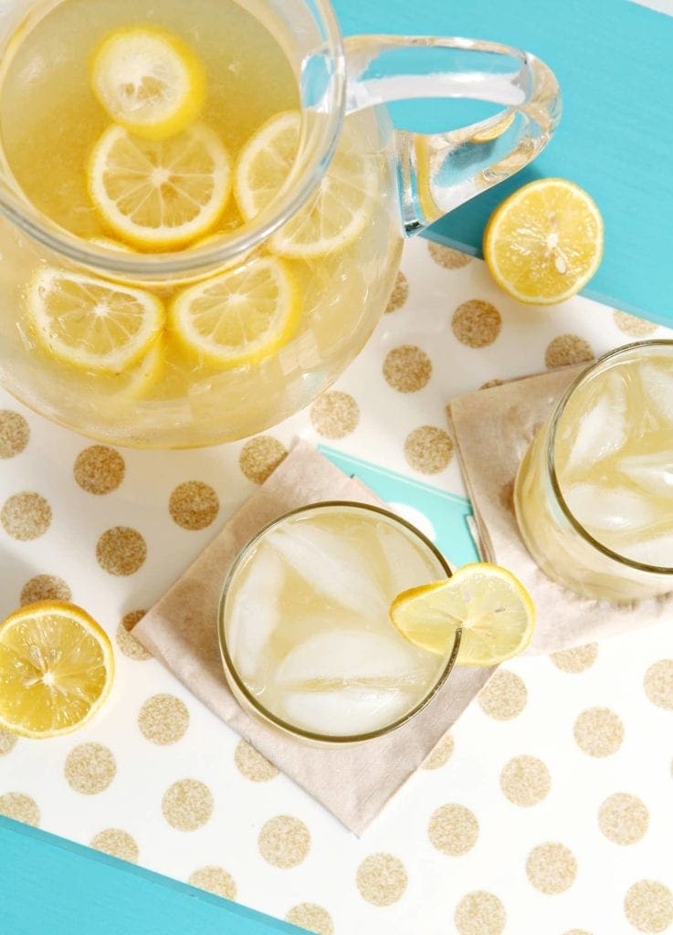 Two glasses of honey lemonade sit on a gold and white polka dot platter next to a pitcher on a bright blue tabletop