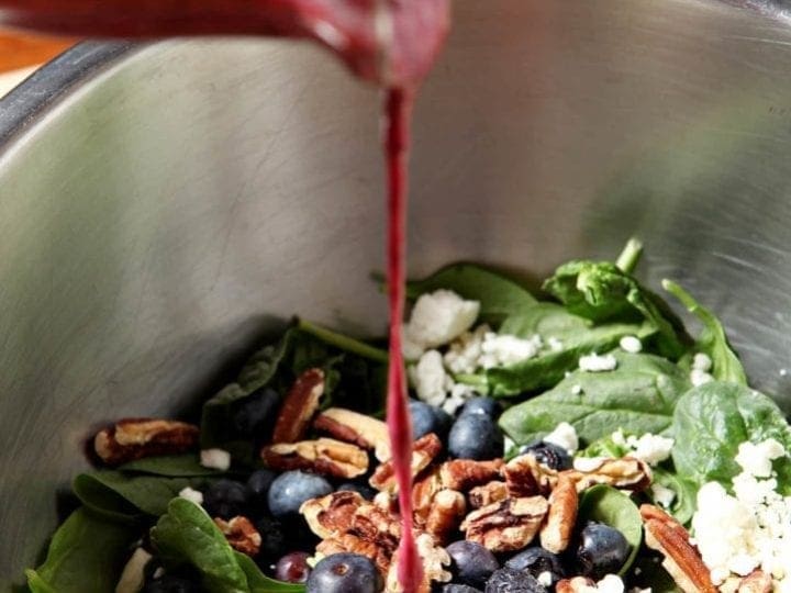 Homemade Blueberry Vinaigrette is poured into a large bowl containing the ingredients for Blueberry Pecan Feta Salad