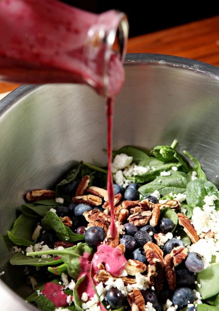 Homemade Blueberry Vinaigrette is poured into a large bowl containing the ingredients for Blueberry Pecan Feta Salad