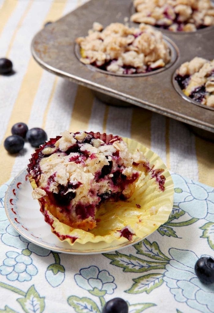 Dairy-Free Blueberry Lemon Crumble Muffin sits on a patterned blue napkin, with additional muffins in a tin and blueberries in the background