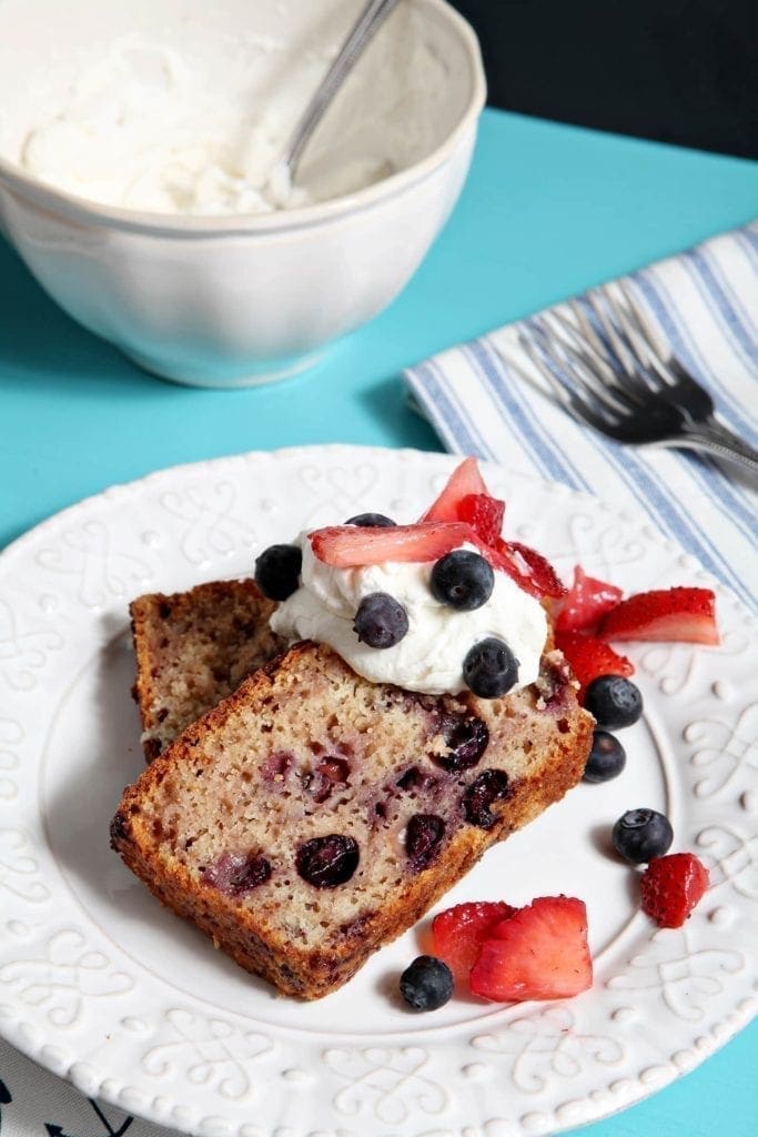 Two slices of Berry Pound Cake, topped with homemade whipped cream and fresh berries, sit on a white plate on a turquoise background