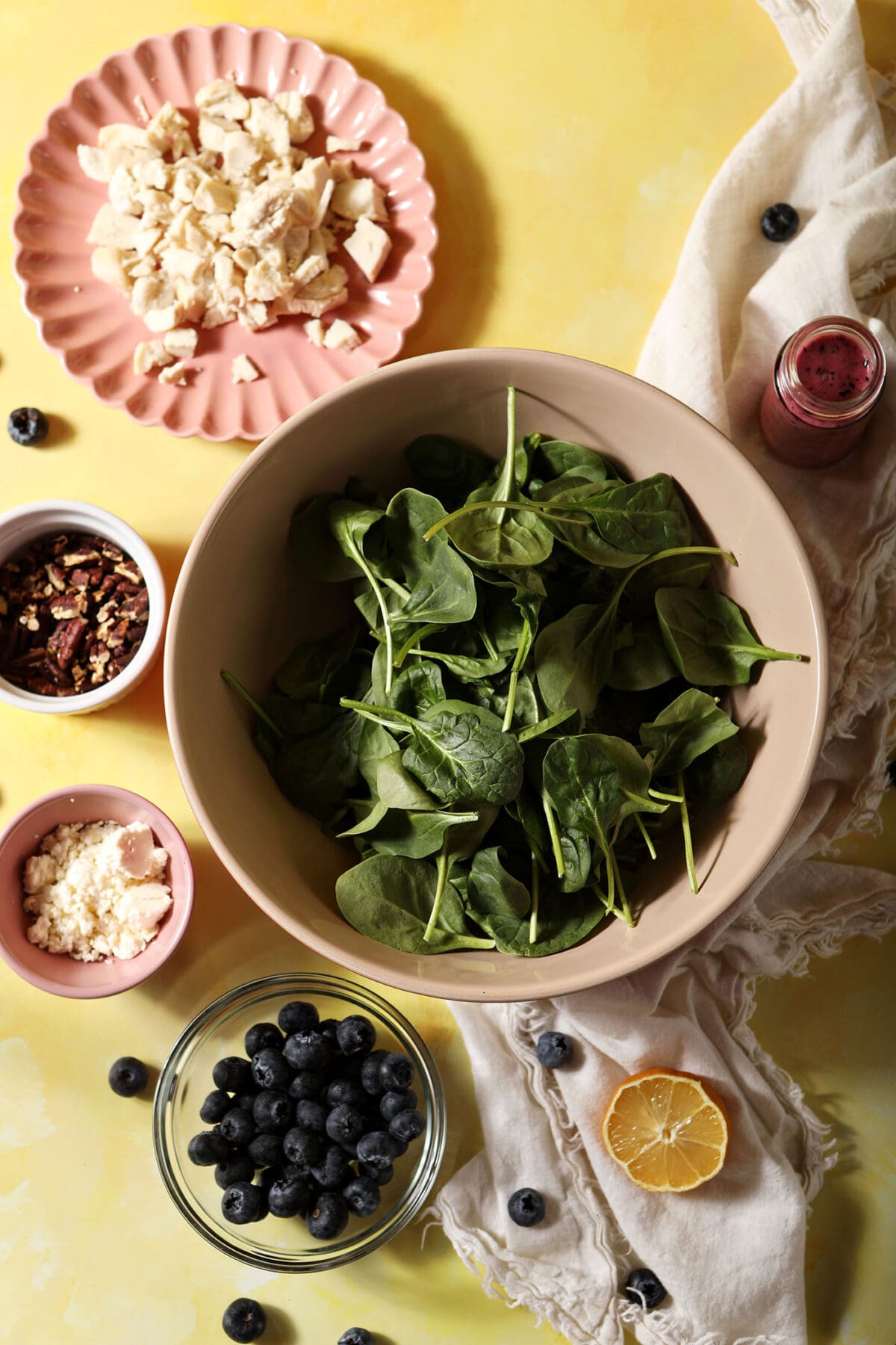 ingredients to make a Blueberry Salad with Feta, Chicken and Pecans in bowls on a yellow surface with a white linen