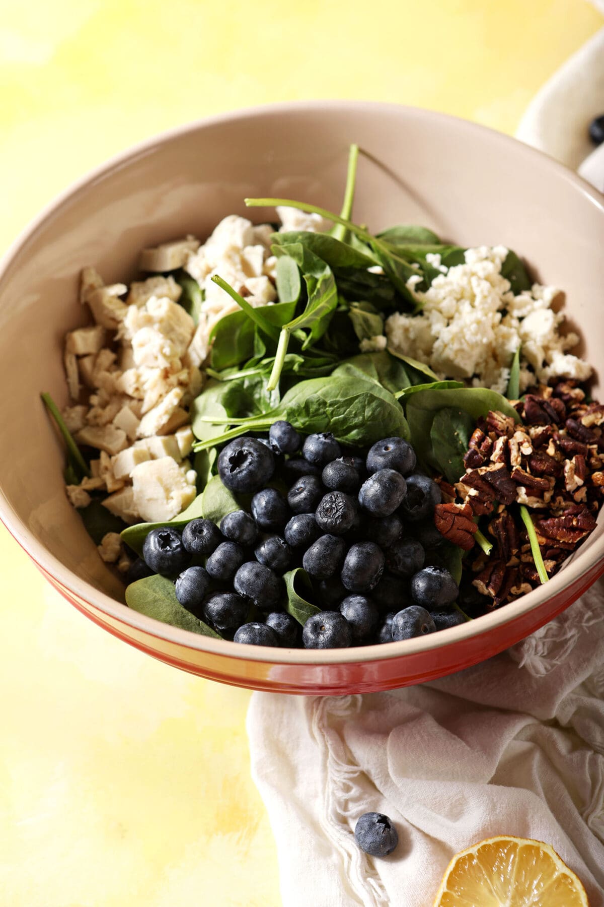 A red bowl holds ingredients to make a blueberry salad, before dressing or tossing, on a yellow surface