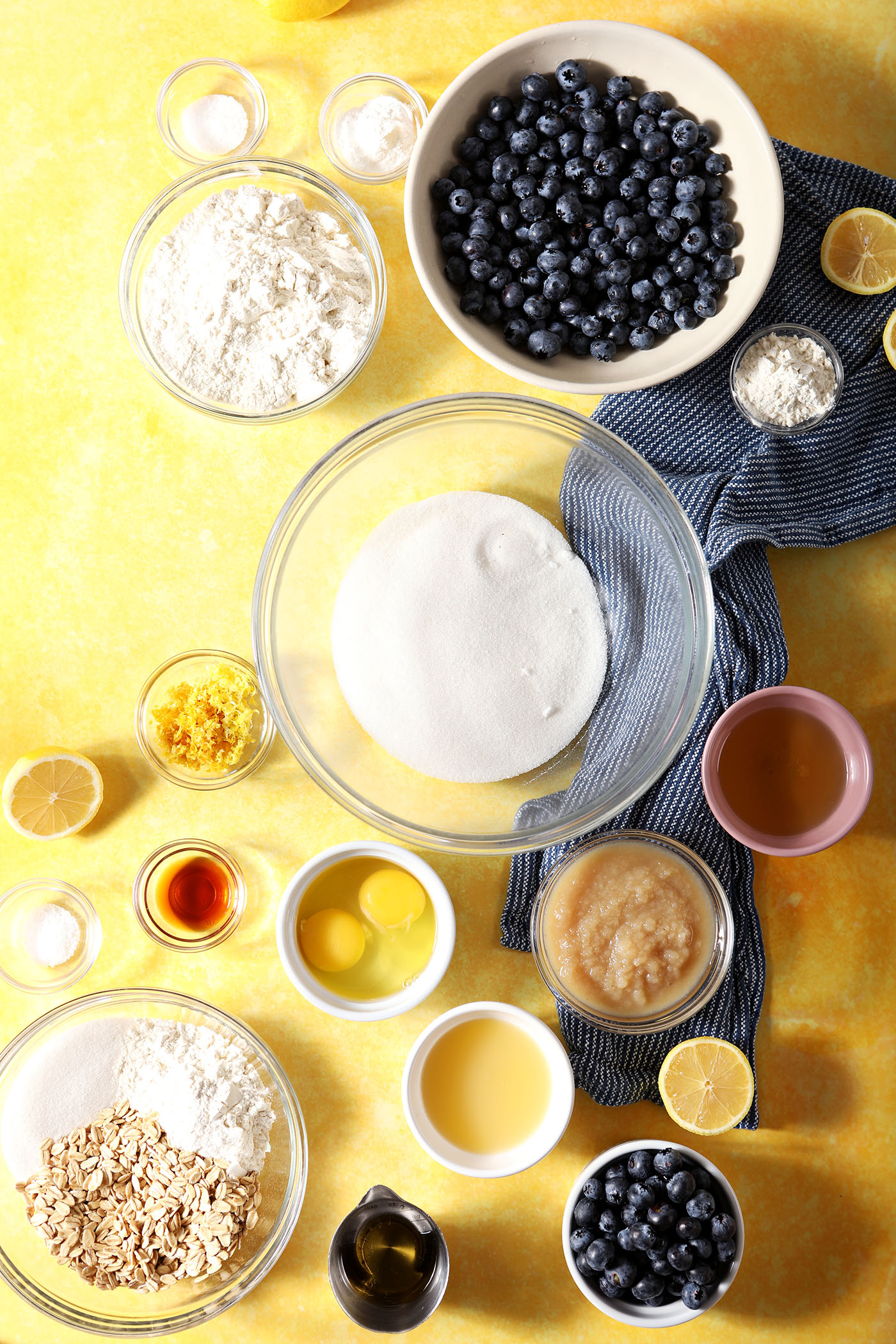 Ingredients to make lemon blueberry muffins with streusel in bowls on a yellow surface with a blue linen