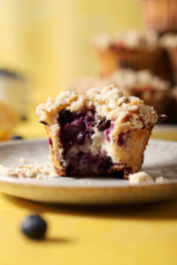 Close up of a bitten-into blueberry lemon muffin with streusel on a grey plate on a yellow surface