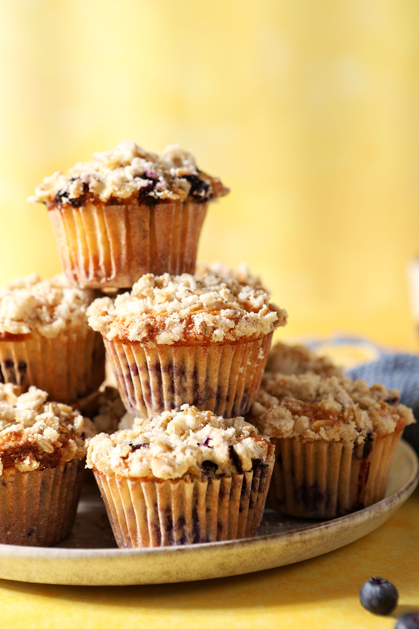 A stack of Lemon Blueberry Muffins with Streusel Topping on a grey plate on a yellow surface near a blue linen