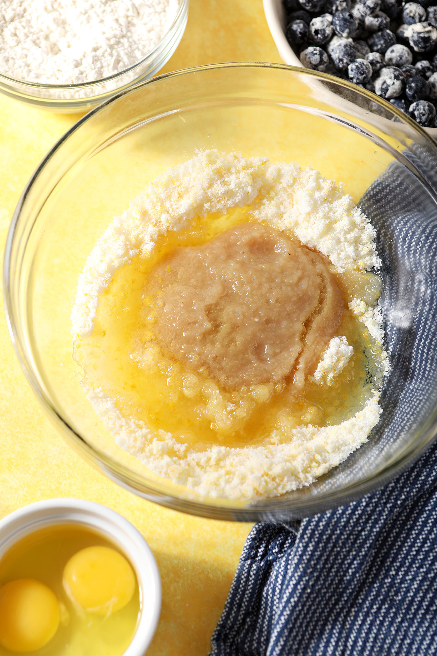 Oil and applesauce on top of lemon sugar in a bowl on a yellow surface next to a blue linen