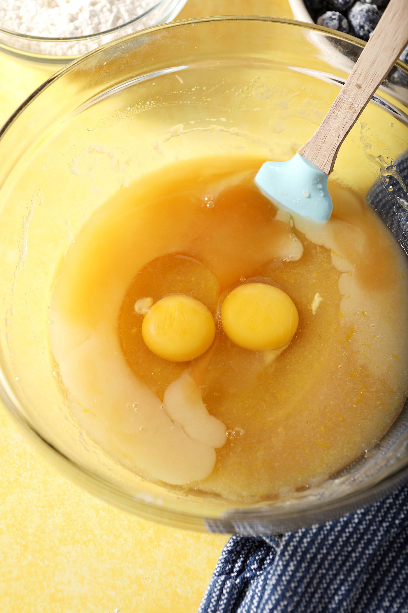 Eggs and vanilla extract on top of other wet ingredients in a bowl on a yellow surface near a blue linen