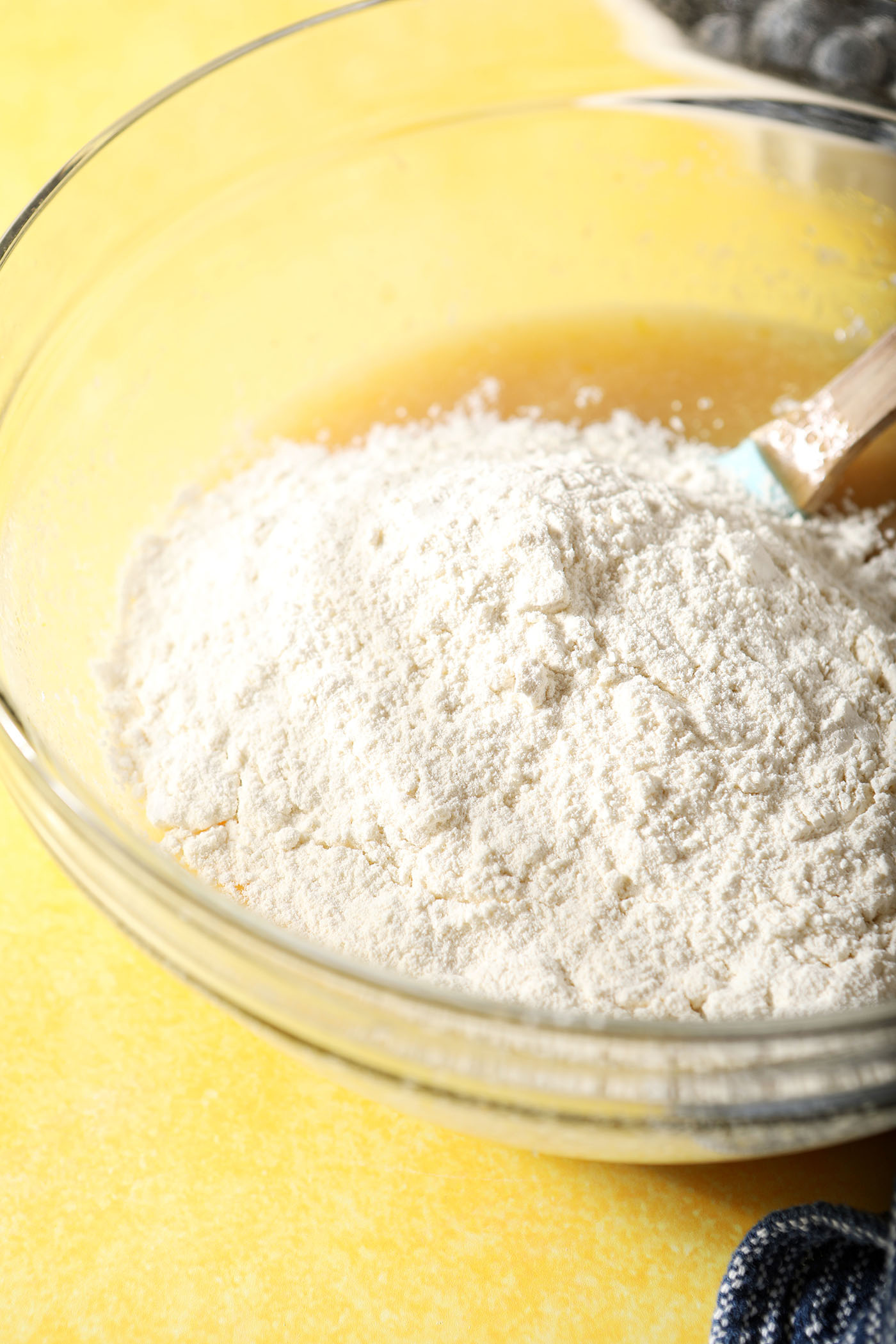 A flour mixture sits on top of wet ingredients in a glass bowl on a yellow surface before mixing