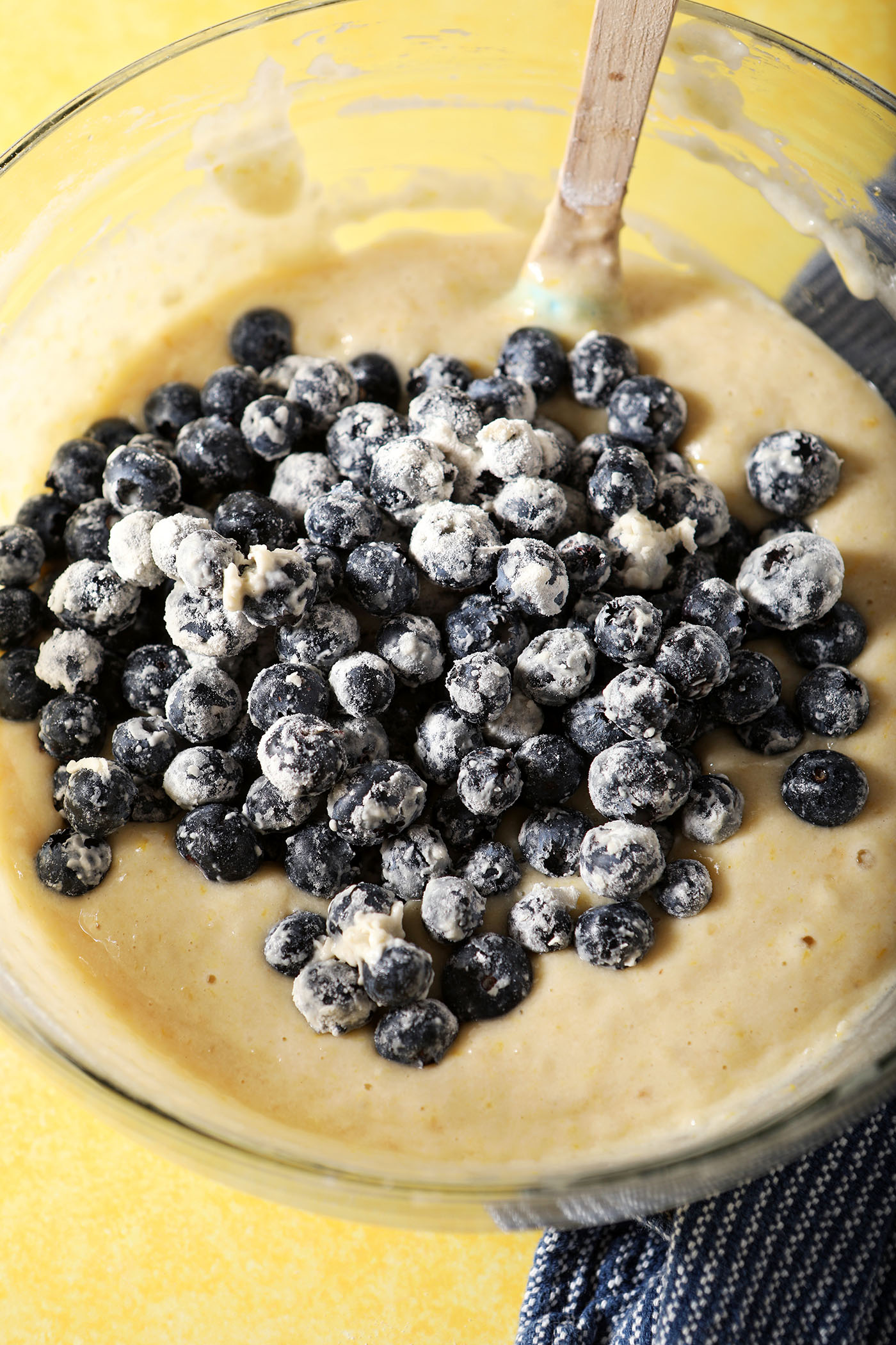 Blueberries coated in flour sit on top of muffin batter in a bowl on a yellow surface