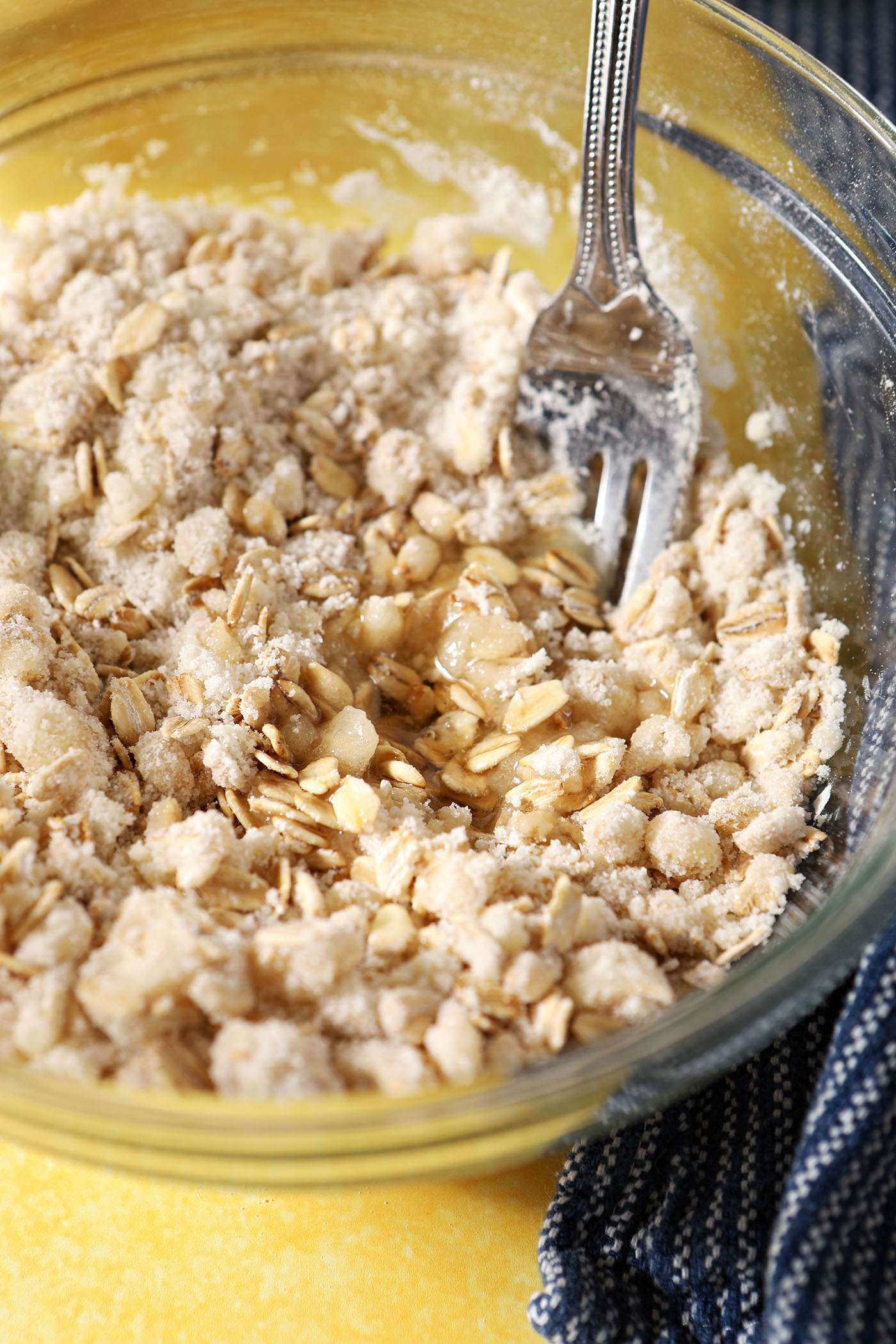 Close up of a crumble topping after mixing in a bowl on a yellow surface next to a blue linen