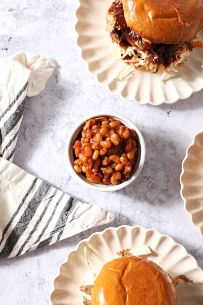 A small ramekin holds a serving of baked beans next to plates of pulled pork sandwiches