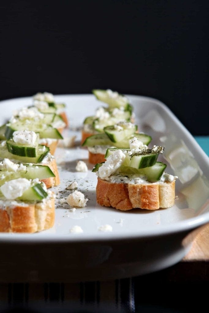 Cucumber Bruschetta, served on a white platter, sits on a wooden background and is ready for enkoying