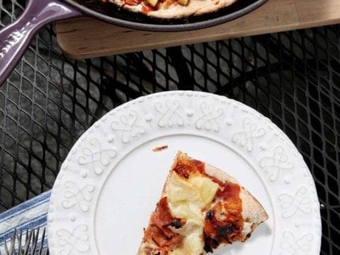 A slice of Grilled Hawaiian Pizza on white plate next to skillet of pizza 