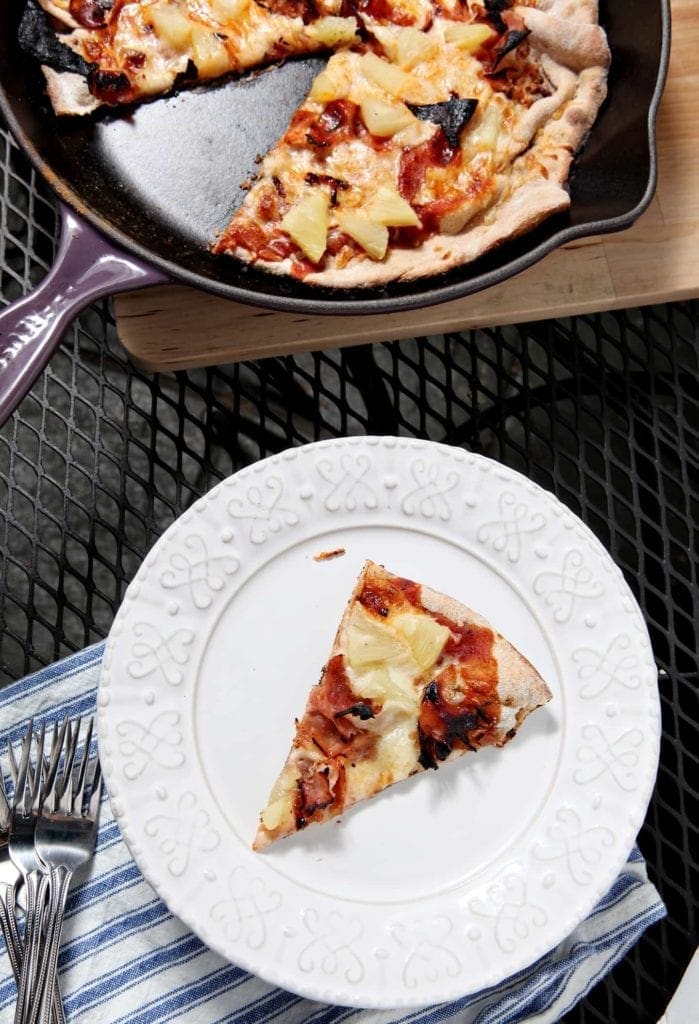 A slice of Grilled Hawaiian Pizza sits on a white platter on an outdoor table with the rest of the pizza, shown in a cast iron skillet.