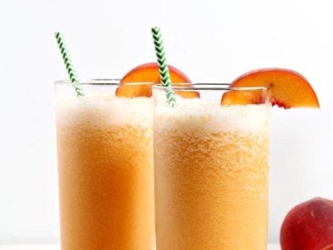 Two Bourbon Peach Slushes, garnished with peach slices and green chevron straws, sit on a marble tile.