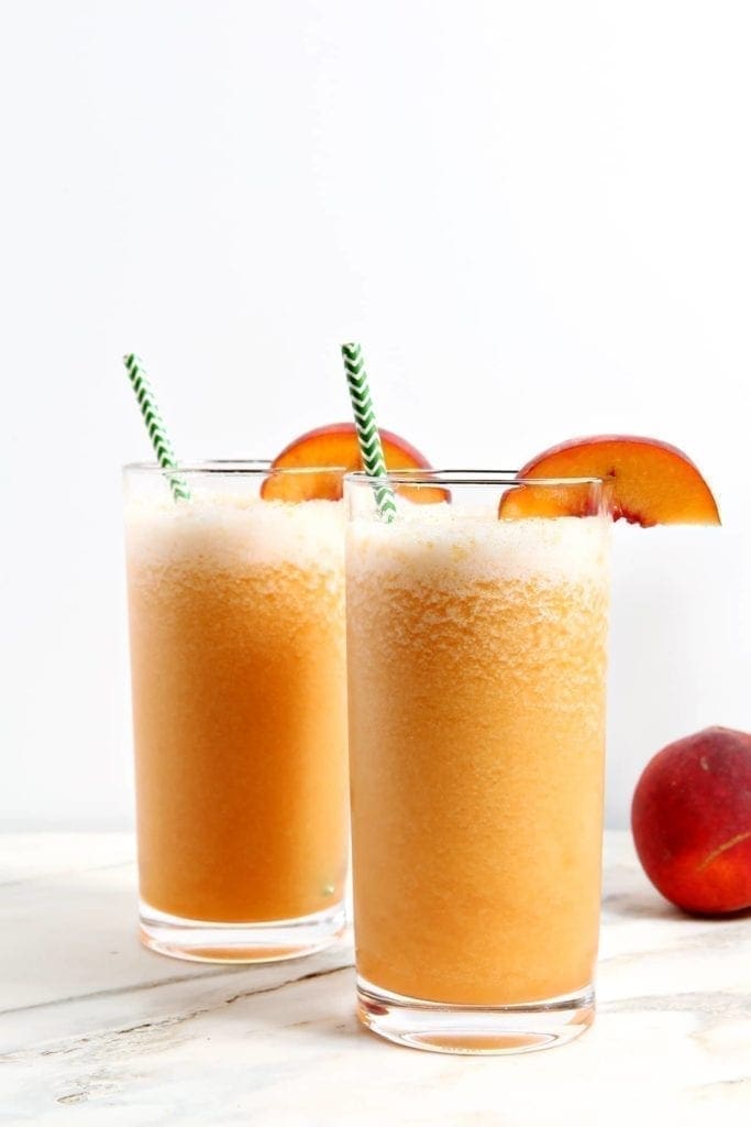 Two Bourbon Peach Slushes, garnished with peach slices and green chevron straws, sit on a marble tile.