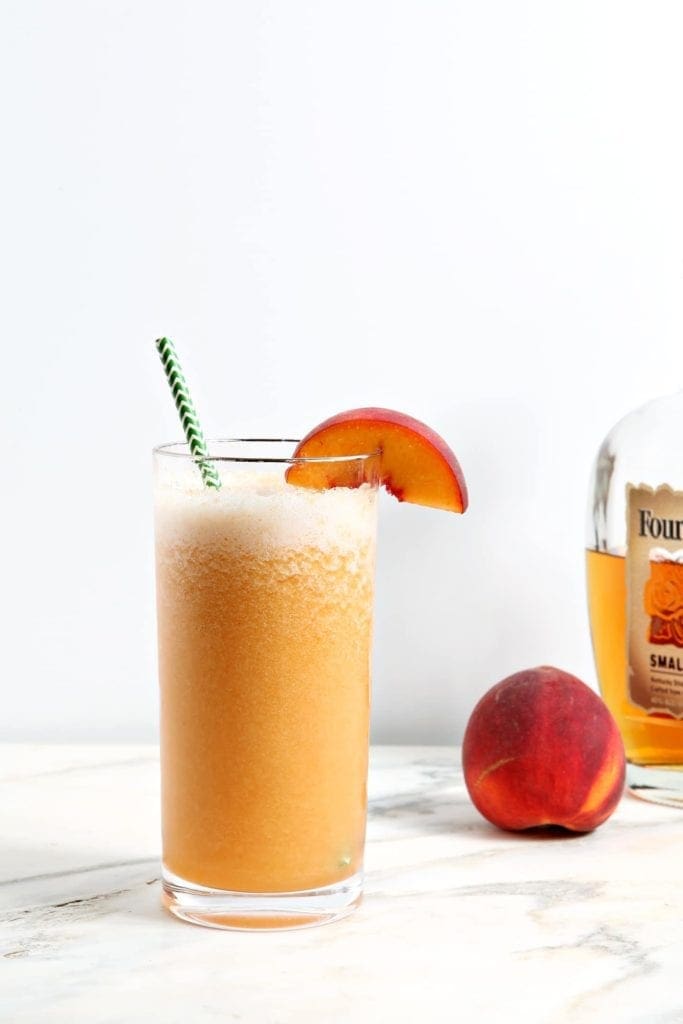 A highball glass holds an orange peach cocktail in it, garnished with a peach