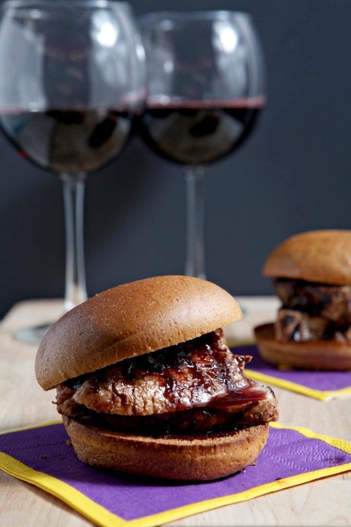 Two Pork Tenderloin Sliders with a Red Wine Reduction Sauce are served with glasses of red wine on a wooden board