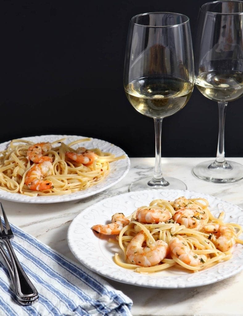 Two white plates hold Lemon Shrimp Pasta on marble with two forks and two glasses of white wine