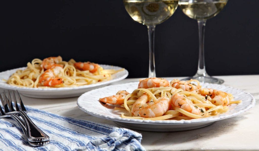 Two white plates of Shrimp Piccata on marble with a blue and white striped towel and two glasses of wine