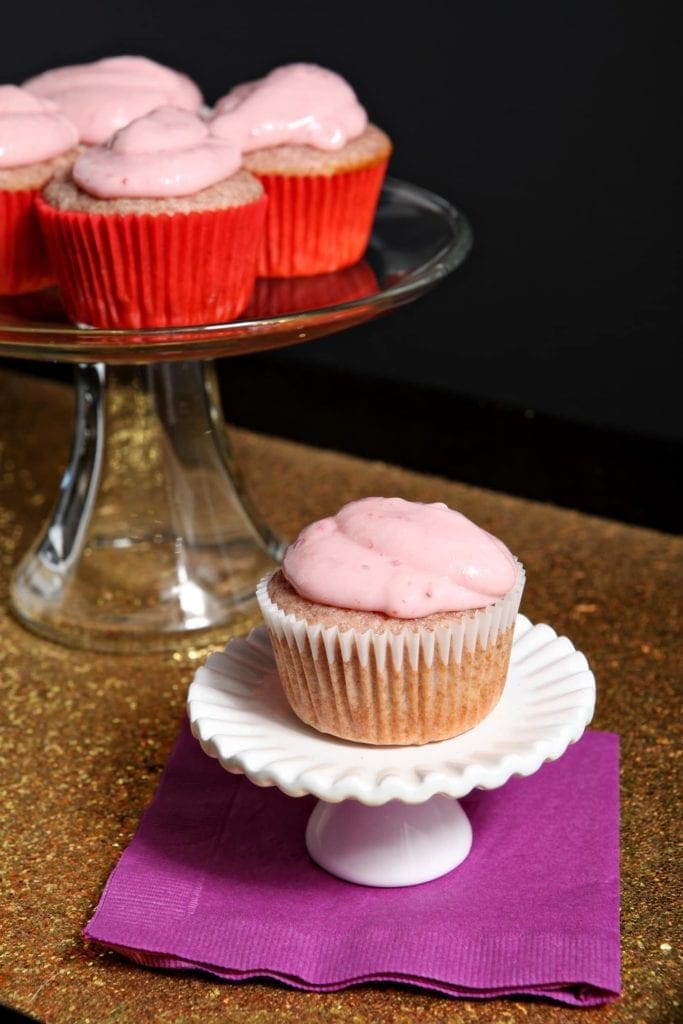 A strawberry cupcake on a stand next to other strawberry cupcakes on a bigger stand on a gold sparkly tray