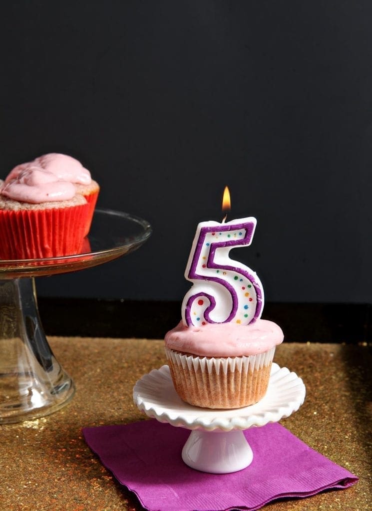 Cupcake sitting on white stand with lit number 5 birthday candle on top 