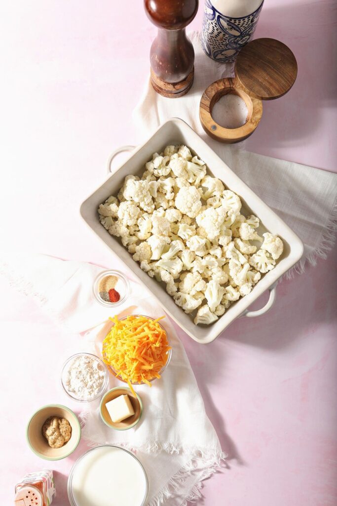 Ingredients to make a cheesy cauliflower casserole in bowls and dishes on a pink countertop