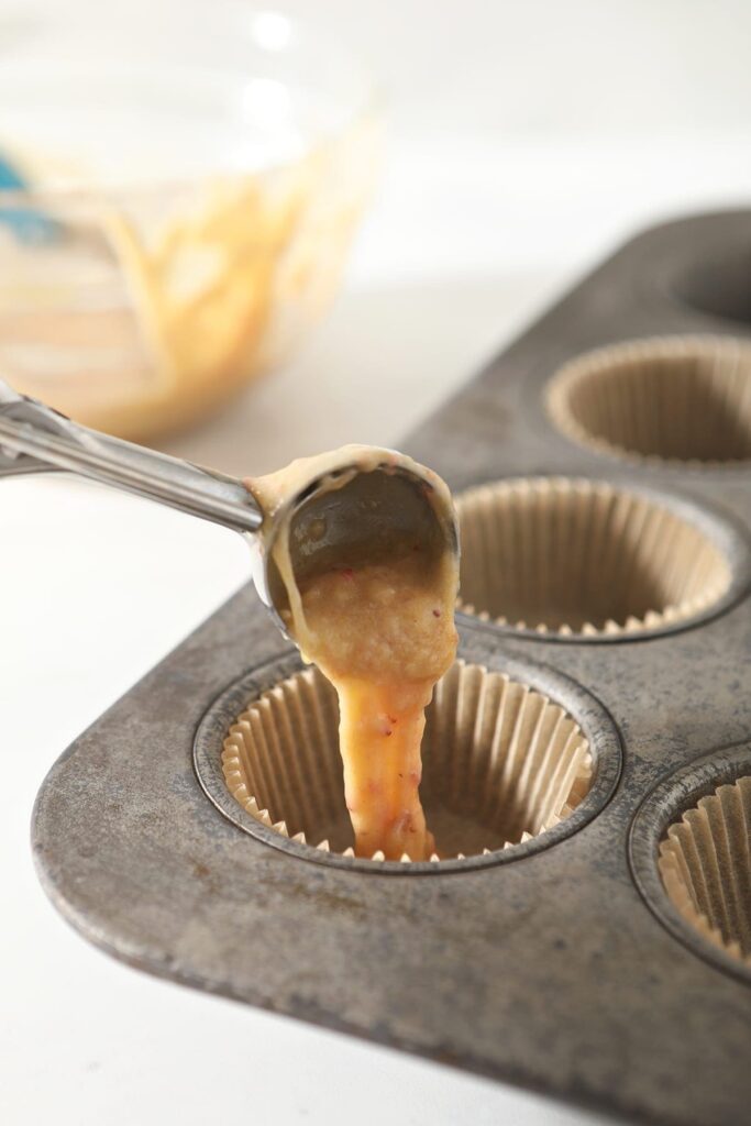 A cookie scoop pours cupcake batter into a muffin tin