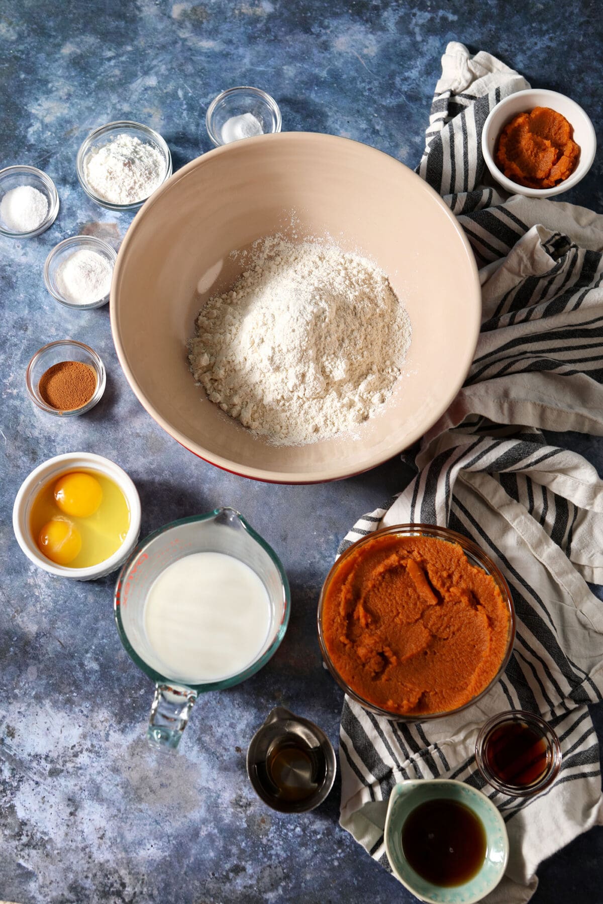 Ingredients to make homemade waffles with pumpkin puree in bowls on a blue surface with white and black striped linens