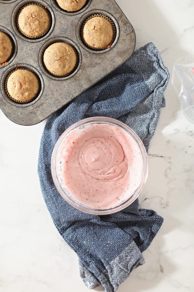 Mixed strawberry cream cheese frosting in a bowl next to baked cupcakes