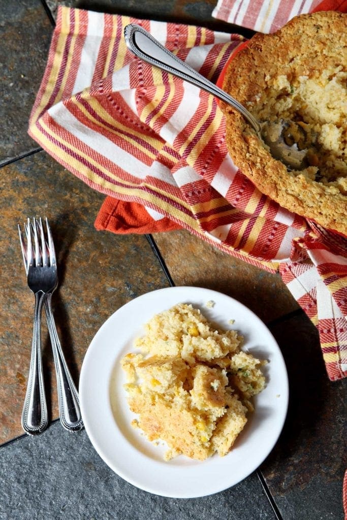 A white plate holds a serving of Creamed Cornbread Casserole on a dark table with forks and a casserole dish