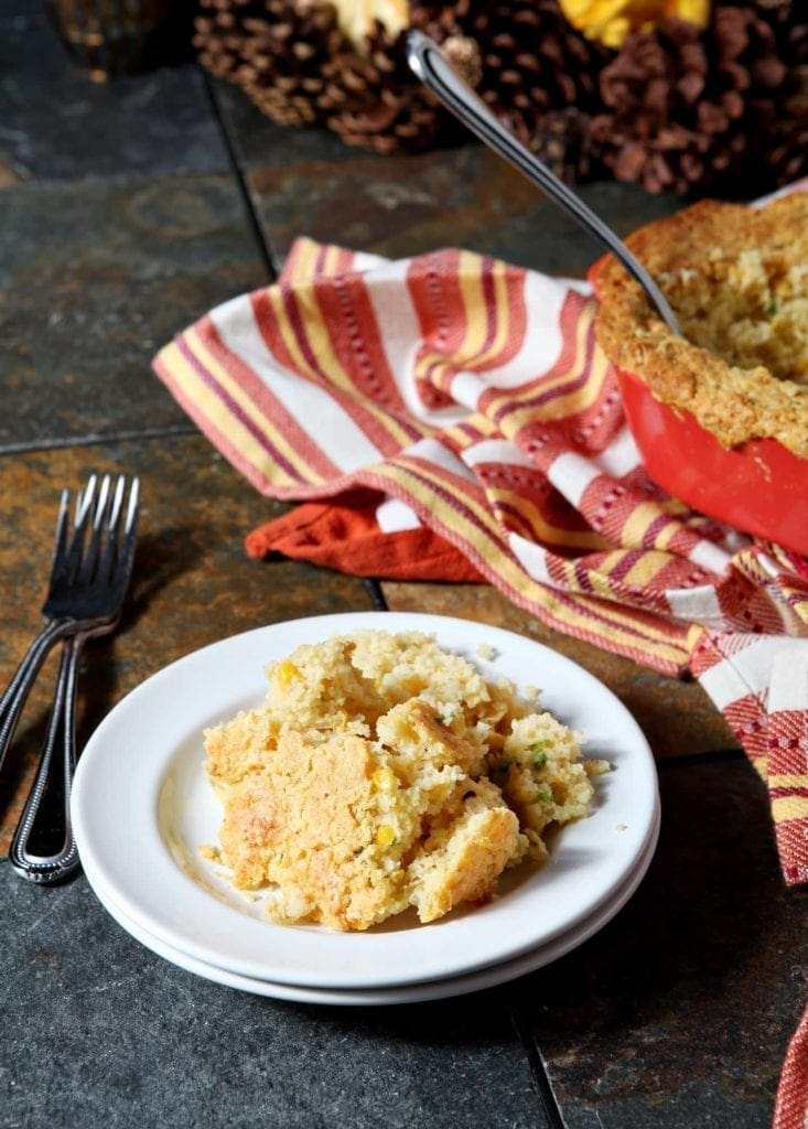 A white plate holds a serving of Creamed Cornbread Casserole, with the casserole dish in the background