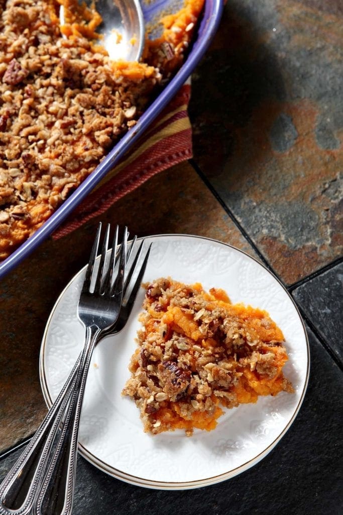 A plate of Bourbon Sweet Potato Casserole sits next to the purple casserole dish holding the Thanksgiving side dish