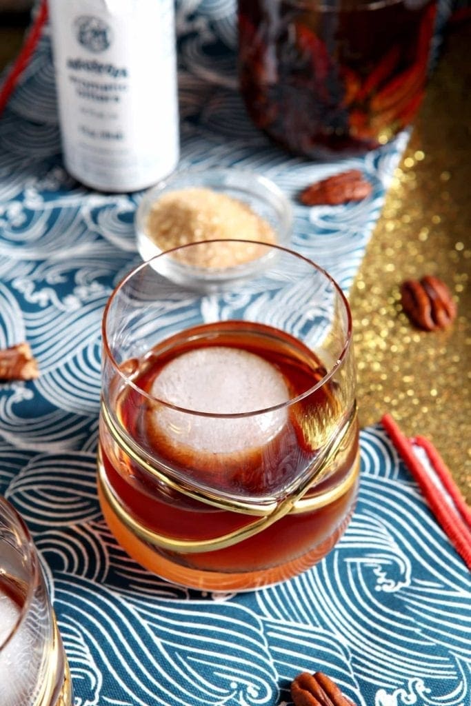 A Toasted Pecan Old Fashioned sits in a fancy glass on a blue and gold background with the ingredients