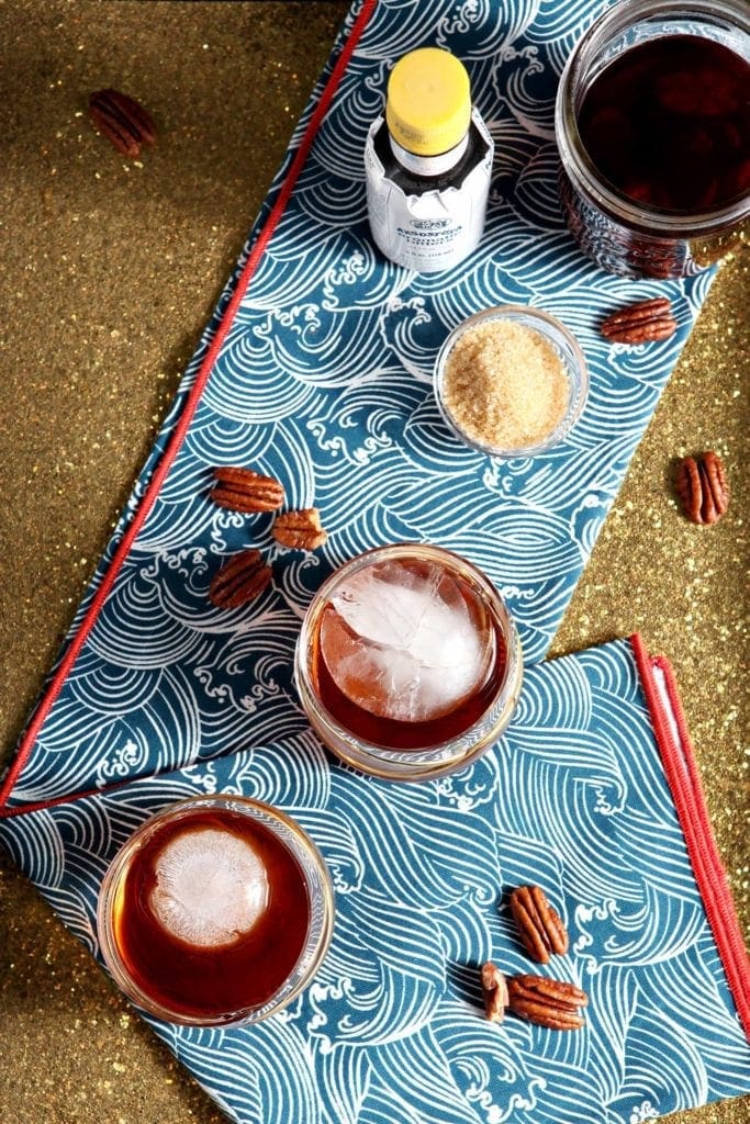 Overhead image of two whiskey cocktails sitting on a blue-and-white dishtowel on a gold glittery tray with pecans, sugar and bitters