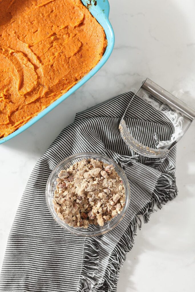 The streusel topping in a bowl next to a casserole dish holding the sweet potato filling
