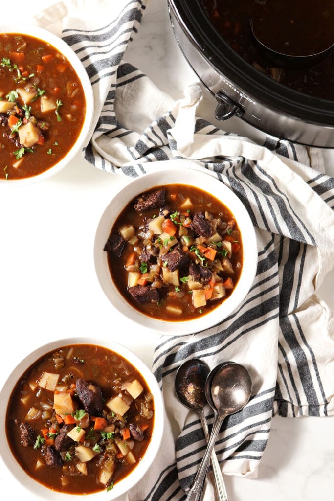 Three bowls of Slow Cooker Beef Stew on a marble surface with blue and white striped towels next to a slow cooker